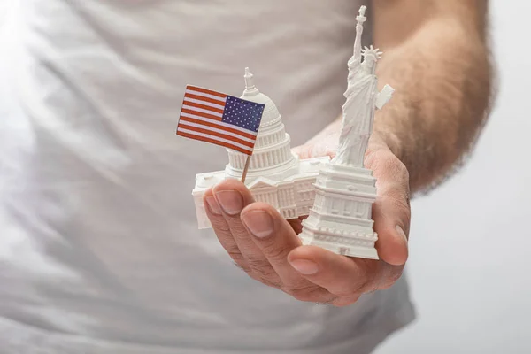 Vista recortada del hombre sosteniendo una pequeña estatua de la libertad y el capitolio de los estados unidos cerca de una pequeña bandera americana aislada en blanco - foto de stock