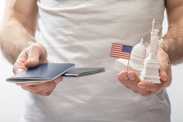 Vista cortada do homem que prende passaportes, estátua pequena da liberdade e capitol dos estados unidos perto da bandeira americana pequena isolada no branco — Fotografia de Stock