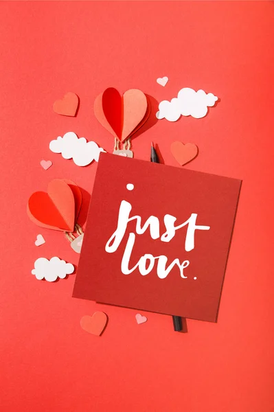 Top view of paper heart shaped air balloons in clouds near card with just love lettering on red background — Stock Photo