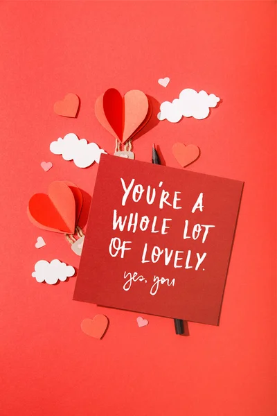 Top view of paper heart shaped air balloons in clouds near card with you're a whole lot of lovely yes, you lettering on red background — Stock Photo