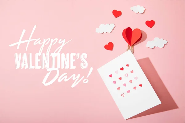 Top view of greeting card with hearts near paper heart shaped air balloon in clouds and  happy valentines day lettering on pink — Stock Photo