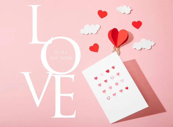 Top view of greeting card with hearts near paper heart shaped air balloon in clouds and love is all you need lettering on pink — Stock Photo