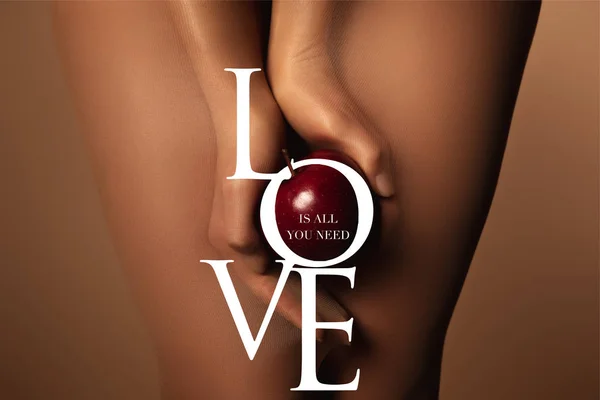 Cropped view of woman in nylon tights holding ripe red apple near love is all you need lettering on brown — Stock Photo