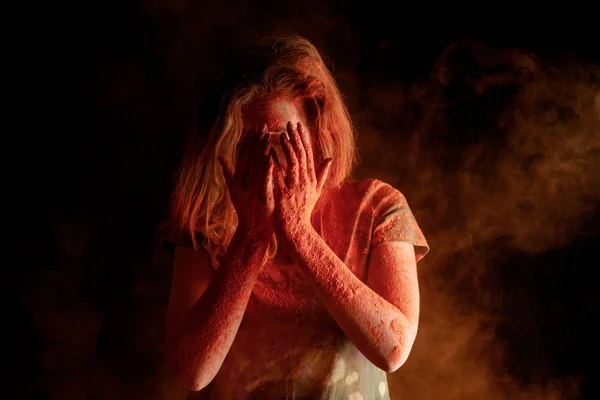 Mujer cubriendo la cara con pintura holi de color naranja en las manos sobre fondo negro - foto de stock