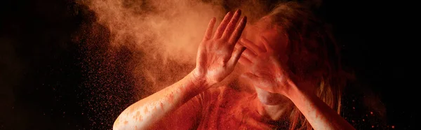 Woman covering face from orange colorful holi paint cloud on black background, panoramic shot — Stock Photo