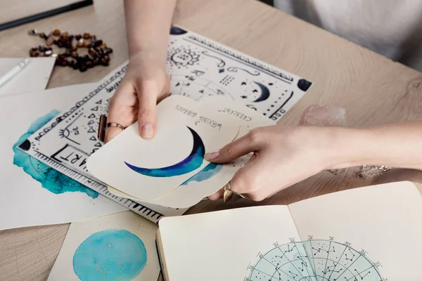 Vista ritagliata dell'astrologo che tiene carte con disegni ad acquerello per tema di nascita con segno zodiacale sul tavolo — Foto stock