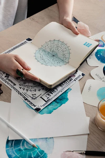 Ausgeschnittene Ansicht eines Astrologen, der ein Notizbuch mit Karten mit Aquarellzeichnungen von Tierkreiszeichen auf dem Tisch hält — Stockfoto