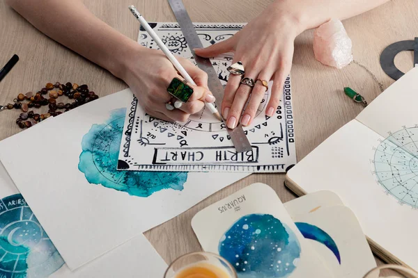 Ausgeschnittene Ansicht eines Astrologen, der Geburtshoroskope mit Aquarellbildern mit Tierkreiszeichen auf Karten auf dem Tisch zeichnet — Stockfoto