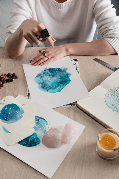 Vista recortada del astrólogo sosteniendo el cristal en la cadena por encima de las pinturas de acuarela con signos del zodíaco en las tarjetas en la mesa - foto de stock