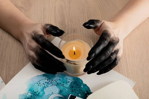 Cropped view of witch with black paint on hands holding candle by cards with watercolor zodiac signs on table — Stock Photo