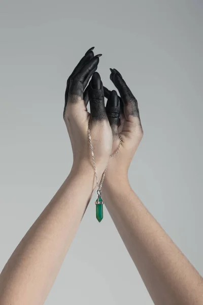 Cropped view of witch with black paint on hands holding green crystal on chain isolated on grey — Stock Photo