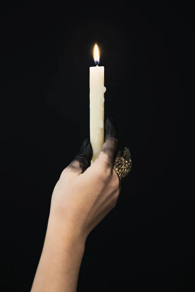 Cropped view of witch with black paint on hand and jewelry ring holding candle isolated on black — Stock Photo
