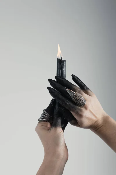 Vista ritagliata di mani di strega con dita dipinte di nero e anelli di gioielli che tengono la candela isolata sul grigio — Foto stock
