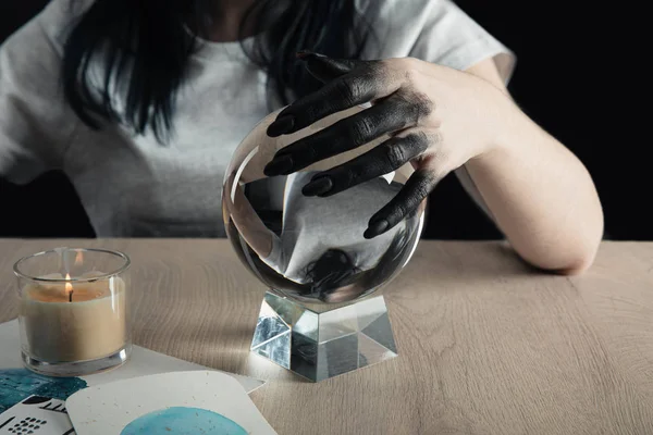 Cropped view of magician with black painted hand holding crystal ball by watercolor paintings and candle on table isolated on black — Stock Photo