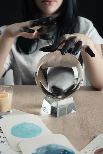 Cropped view of magician with black paint on hands, crystal ball and watercolor paintings on table isolated on black — Stock Photo