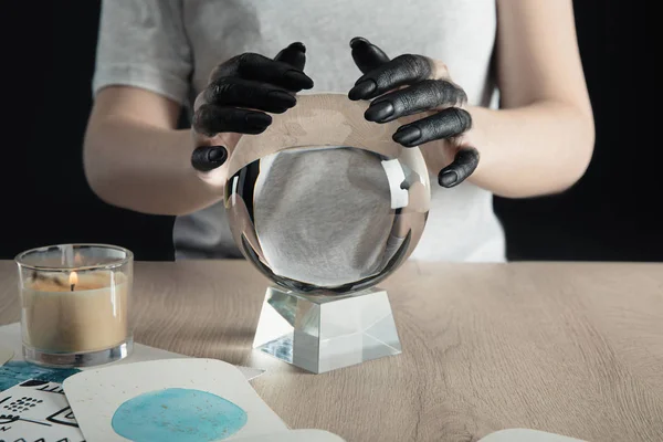 Vista ritagliata della strega con le mani tinte di nero che tengono palla di cristallo da candela e disegni ad acquerello sul tavolo isolato su nero — Foto stock