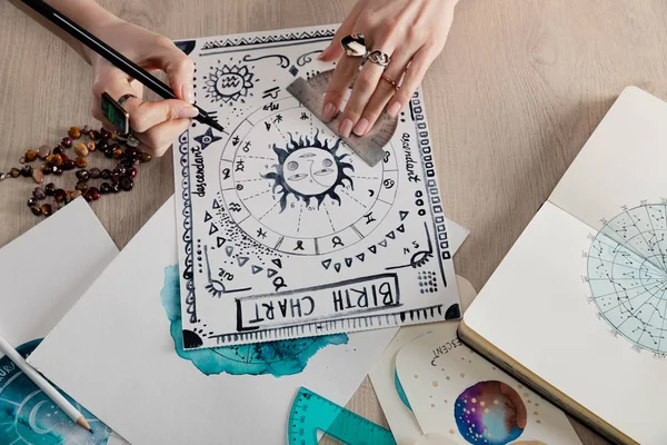 Vista de ángulo alto de las pinturas de acuarela con signos del zodiaco en las tarjetas y carta natal de dibujo astrólogo en la mesa - foto de stock