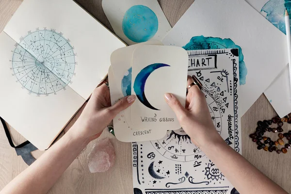 Top view of astrologer holding watercolor paintings with moon phases on cards by birth chart on table — Stock Photo