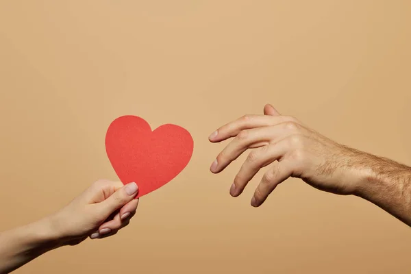 Vista recortada de la mujer sosteniendo el corazón rojo y el hombre alcanzándolo aislado en beige - foto de stock