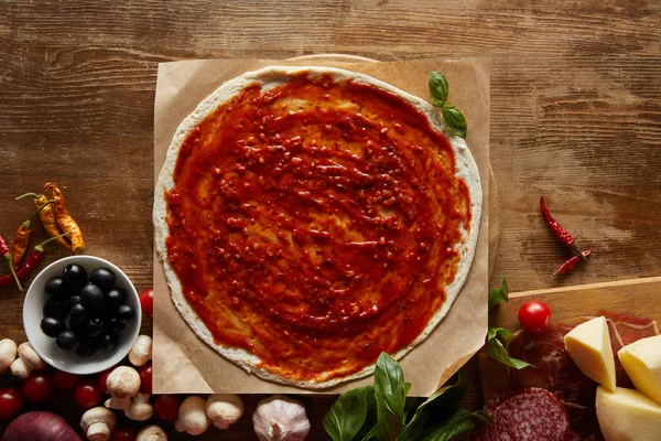 Top view of pizza on parchment paper with basil, vegetables, olives, prosciutto, salami, mushrooms and cheese on wooden background — Stock Photo