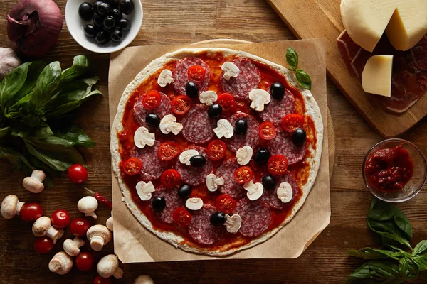 Vista superior de pizza con salami, champiñones, aceitunas, salsa de tomate, verduras, parmesano y jamón sobre fondo de madera — Stock Photo