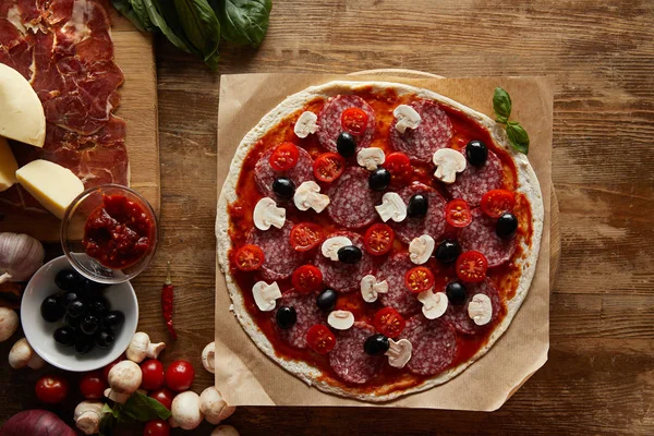 Vista superior de pizza con salami, champiñones, aceitunas, salsa de tomate, parmesano, tomates cherry y jamón sobre fondo de madera - foto de stock