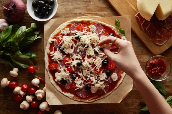 Cropped view of woman making pizza with olives, cherry tomatoes and salami on wooden background — Stock Photo