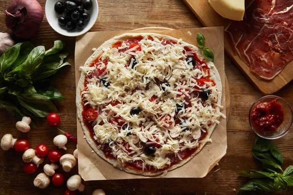 Vista superior de la pizza sobre papel pergamino con parmesano, verduras, aceitunas y salami sobre fondo de madera - foto de stock