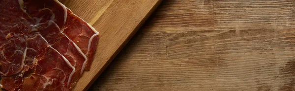 Vista superior del jamón en rodajas sobre tabla de cortar sobre fondo de madera, plano panorámico - foto de stock