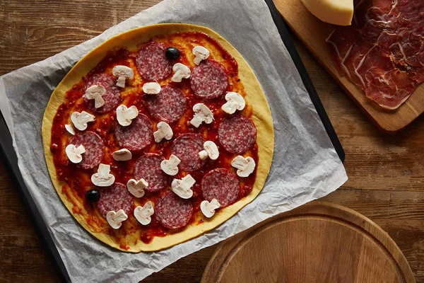 Vista superior de la sabrosa pizza con salami y champiñones sobre papel pergamino sobre fondo de madera - foto de stock