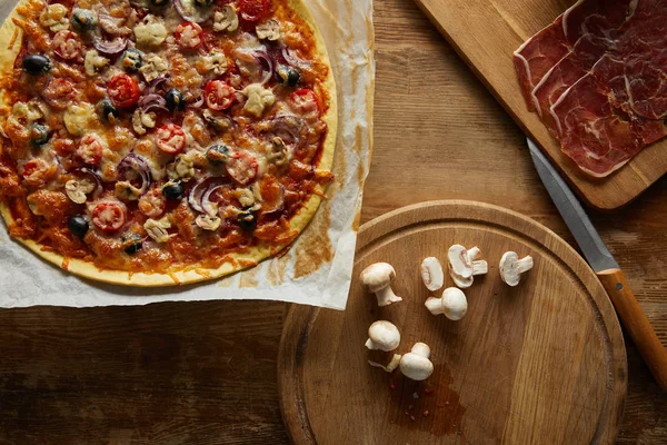 Vista superior de deliciosa pizza sobre papel pergamino, champiñones, jamón y cuchillo sobre fondo de madera - foto de stock