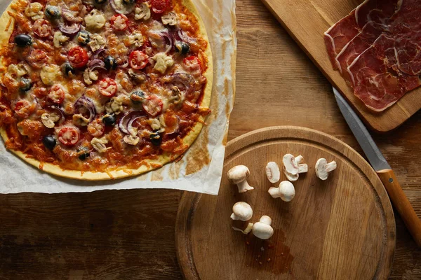 Top view of delicious pizza on parchment paper, mushrooms, prosciutto and knife on wooden background — Stock Photo