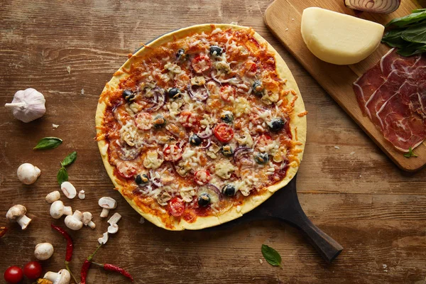 Top view of tasty pizza and garlic, prosciutto, mushrooms, parmesan, chili peppers and basil on wooden background — Stock Photo