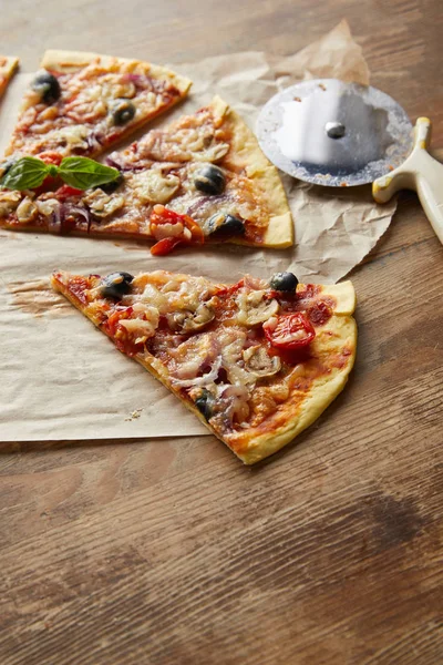 Couper de délicieuses pizzas italiennes aux olives sur papier cuisson avec couteau à pizza sur table en bois — Photo de stock