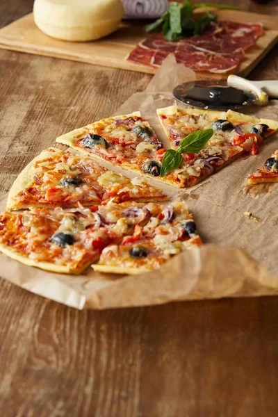 Selektiver Fokus von geschnittener leckerer italienischer Pizza mit Oliven auf Backpapier in der Nähe von Zutaten und Pizzamesser auf Holztisch — Stockfoto