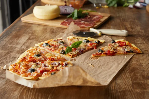 Selektiver Fokus von köstlicher italienischer Pizza mit Oliven auf Backpapier in der Nähe von Zutaten und Pizzamesser auf Holztisch — Stockfoto