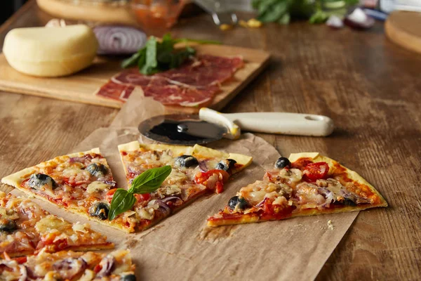Вибірковий фокус на вирізанні смачної італійської піци з оливками на папері для випічки поблизу інгредієнтів та ножем для піци на дерев'яному столі — стокове фото