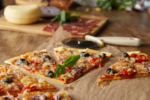 Selektiver Fokus von geschnittener leckerer italienischer Pizza mit Oliven auf Backpapier in der Nähe von Zutaten und Pizzamesser auf Holztisch — Stockfoto