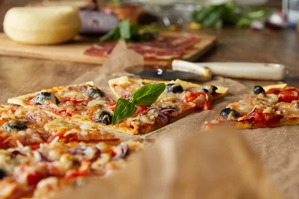 Foco seletivo de corte deliciosa pizza italiana com azeitonas em papel manteiga perto de ingredientes e faca de pizza na mesa de madeira — Fotografia de Stock