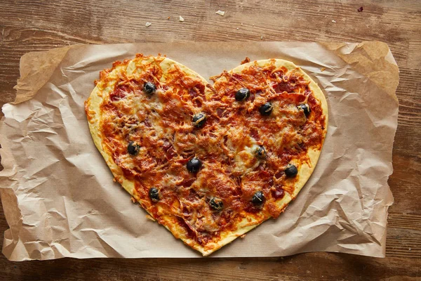 Draufsicht auf köstliche italienische Pizza in Herzform auf Backpapier auf Holztisch — Stockfoto