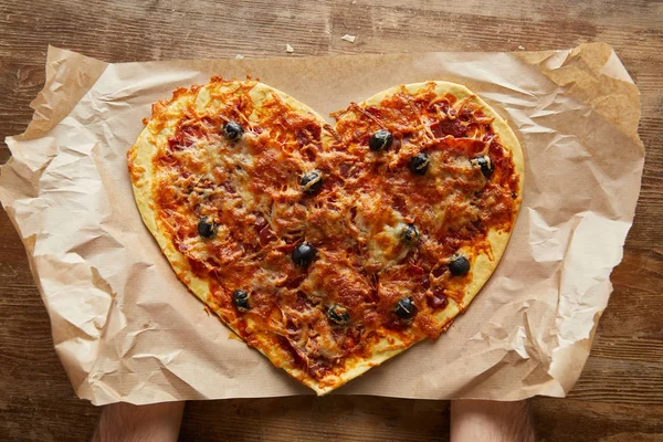 Vista ritagliata dell'uomo che tiene deliziosa pizza italiana a forma di cuore su carta da forno su tavolo di legno — Foto stock