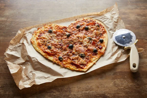 Deliziosa pizza italiana a forma di cuore su carta da forno vicino al coltello da pizza sul tavolo di legno — Foto stock