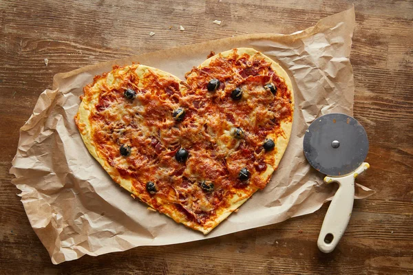 Vista superior da deliciosa pizza italiana em forma de coração no papel manteiga perto de faca de pizza na mesa de madeira — Fotografia de Stock