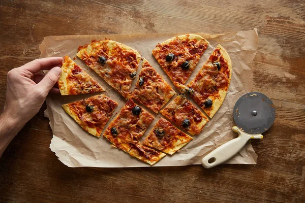 Vista recortada del hombre comiendo deliciosa pizza italiana en forma de corazón cortada en pedazos en papel de hornear cerca de cuchillo de pizza en mesa de madera - foto de stock