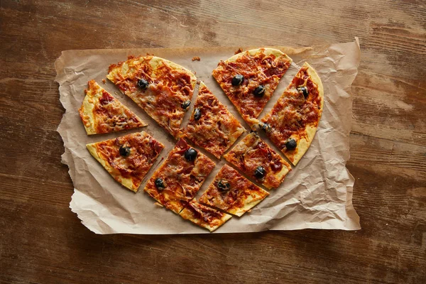 Top view of delicious italian pizza in heart shape cut into pieces on baking paper on wooden table — Stock Photo