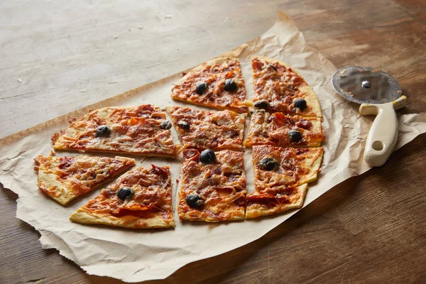 Délicieuse pizza italienne en forme de coeur coupée en morceaux sur du papier cuisson près d'un couteau à pizza sur une table en bois — Photo de stock