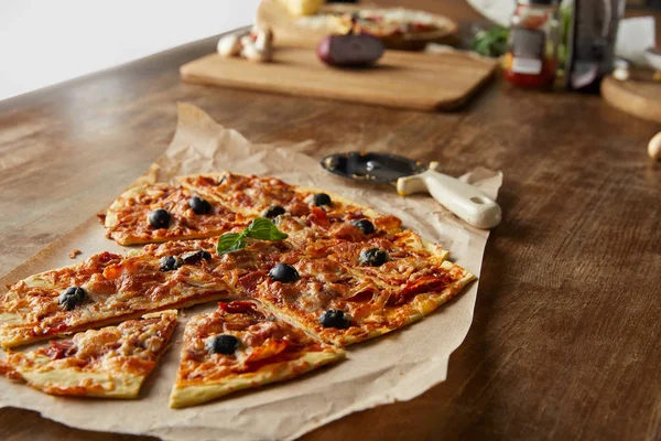 Deliciosa pizza italiana em forma de coração cortada em pedaços em papel manteiga perto de faca de pizza e ingredientes em mesa de madeira — Fotografia de Stock