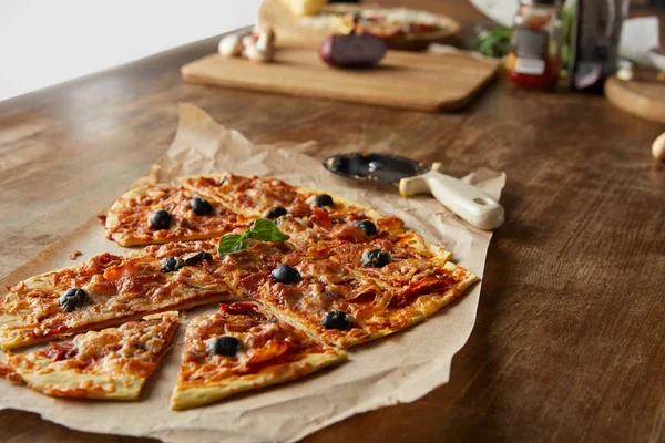 Delicious italian pizza in heart shape cut into pieces on baking paper near pizza knife and ingredients on wooden table — Stock Photo