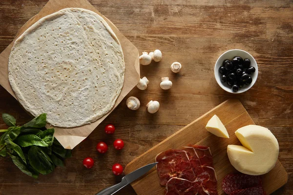 Ansicht von rohem Teig, Kirschtomaten, Basilikum, Pilzen, Oliven, Mozzarella, Salami und Prosciutto für Pizza auf Holztisch — Stockfoto