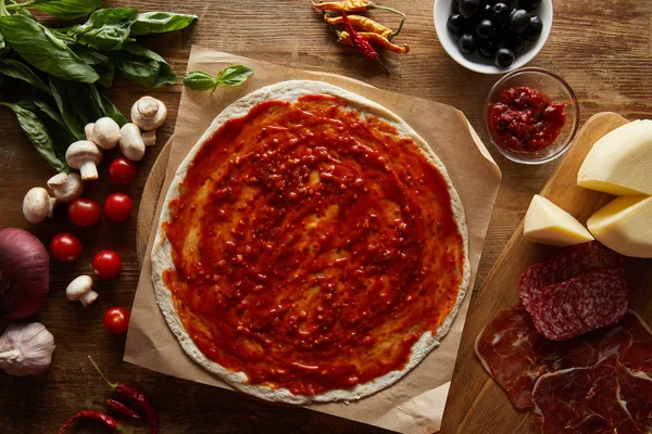 Top view of dough with tomato sauce near ingredients for pizza on wooden table — Stock Photo